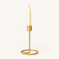 Small gold taper candlestick with a lit ivory taper candle on a warm off-white background
