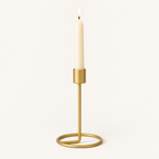 Small gold taper candlestick with a lit ivory taper candle on a warm off-white background