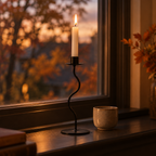 Small wavy iron taper candle holder lit on a windowsill at dusk with autumn trees outside
