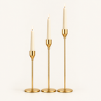 Set of 3 gold taper candlesticks in graduated heights with lit ivory candles on a warm off-white background